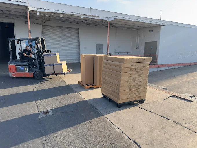 Sideways shot image of two pallets of boxes outside on asphalt with a red forklift moving more boxes towards the pallets.