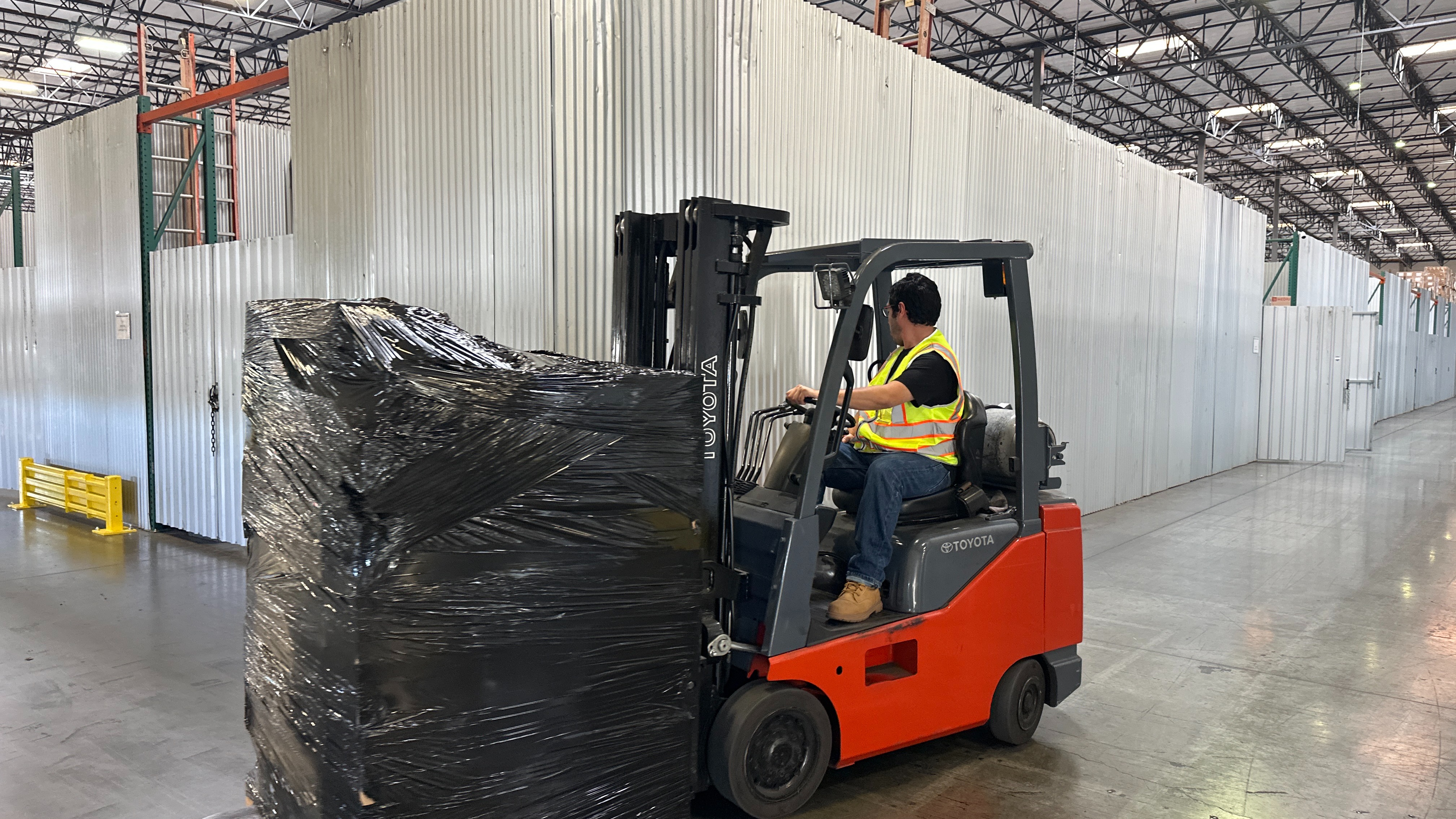 Side shot image of a red forklift with a pallet of products being driven by a man in a yellow vest.